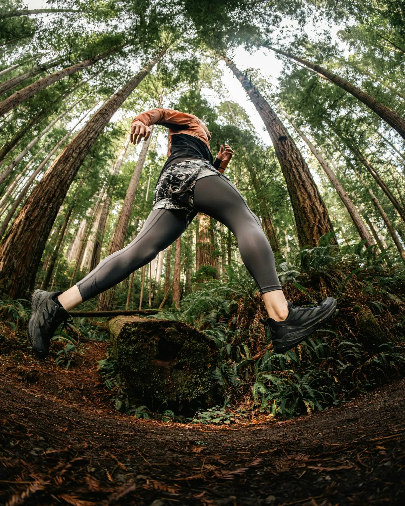 A trail runner mid-stride among redwoods, seen from the ground.